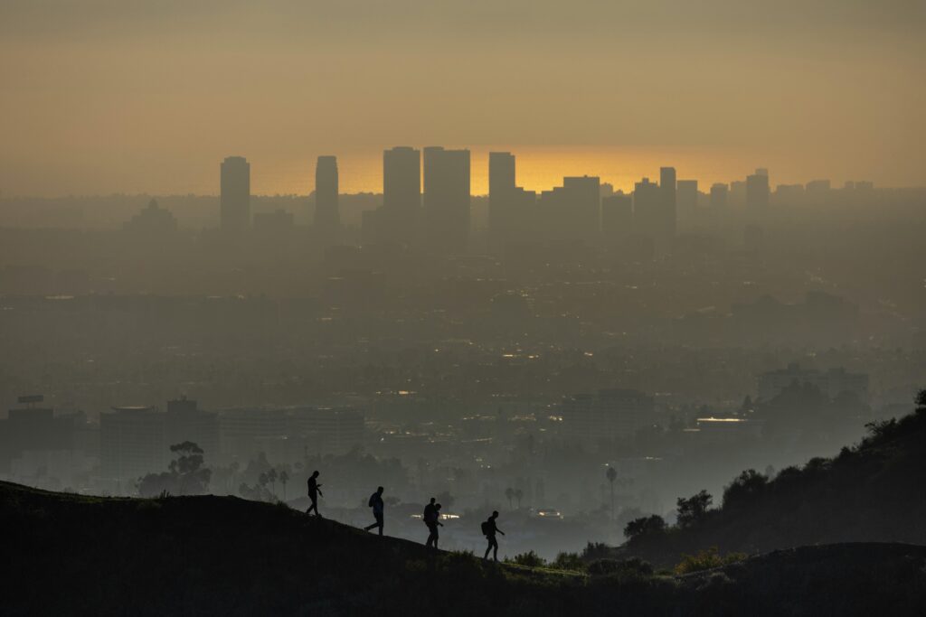 pexels-foto-19774216-19774216 Silhouetted hikers traverse a hill, overlooking the smoggy Los Angeles skyline at sunset.